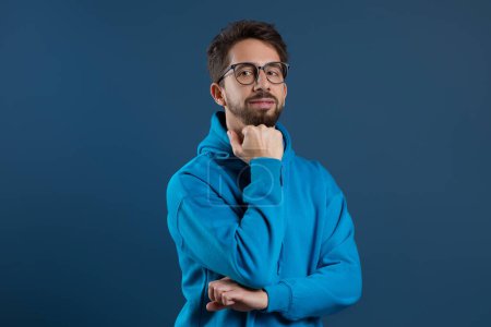 Photo for Pensive young man wearing eyeglasses resting chin on hand and looking at camera, smiling millennial guy wearing blue hoodie looking at camera thoughtfully, standing against dark studio background - Royalty Free Image