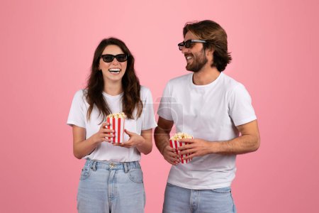 Photo for Laughing european woman and man wearing 3D glasses and holding red and white striped popcorn boxes, enjoying a movie night together, in white t-shirts on a pink background - Royalty Free Image