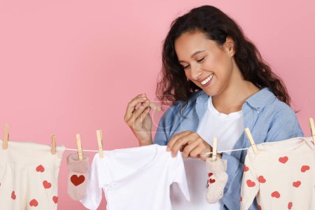 Photo for Joyful motherhood concept. Smiling positive attractive lady young mother putting baby clothes on drier after washing, taking care of her child, pink studio background, copy space - Royalty Free Image