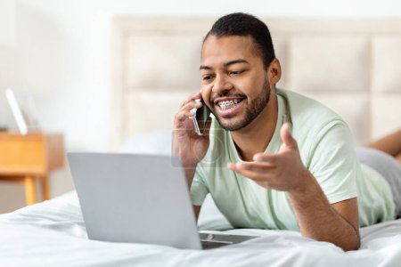 Photo for Cheerful friendly african american man working from home, lying on bed, wearing pajamas, using pc laptop, have phone conversation with assistant or business partner, copy space. Modern business - Royalty Free Image