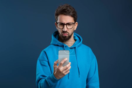 Photo for Skeptical man wearing glasses looking at smartphone screen, young male frowning slightly as if reading something perplexing or disappointing, standing against dark blue studio background, free space - Royalty Free Image
