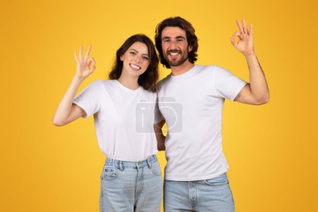 Photo for Joyful european man and woman in white t-shirts and jeans making the OK hand gesture, smiling confidently with a warm yellow background, symbolizing satisfaction and positivity - Royalty Free Image
