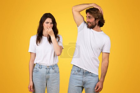 Téléchargez les photos : Une femme européenne se pince le nez d'une expression dégoûtante tandis qu'un homme se gratte la tête avec un sourire, portant à la fois des t-shirts blancs et un jean sur un fond jaune vif - en image libre de droit