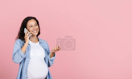 Happy young pregnant woman talking to her expecting friend on phone, gesturing and smiling, looking at copy space isolated on pink studio background, sharing pregnancy experience