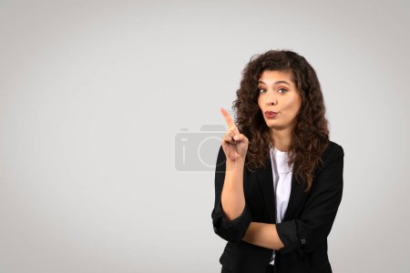 Photo for Engaged young businesswoman with curly hair making pointing gesture, showing free space, looking curious and interested, against clean grey background - Royalty Free Image