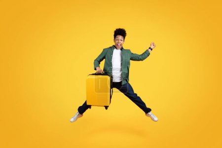 Foto de Emocionado feliz millennial afroamericano chico viajero saltando con la maleta en sus manos, hombre negro turista ir de vacaciones, aislado en el fondo del estudio amarillo. Turismo, oferta caliente - Imagen libre de derechos