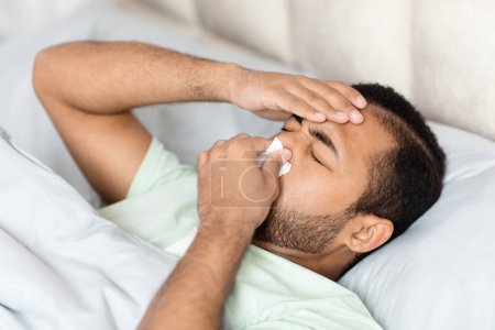 Téléchargez les photos : Malade homme afro-américain millénaire en mauvaise santé portant un pyjama couché au lit, touchant la tête, éternuant, souffrant de rhume, grippe, maux de tête, écoulement nasal, coronavirus, vue grand angle, espace de copie - en image libre de droit