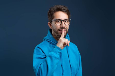 Photo for Make Silence. Portrait Of Young Man Showing Shh Gesture At Camera, Millennia Guy In Eyeglasses Holding Finger Near Lips, Gesturing Hush While Standing Isolated On Blue Studio Background - Royalty Free Image