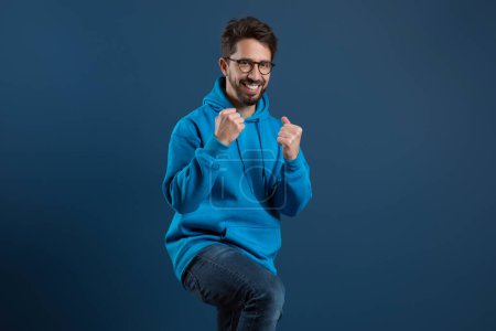 Photo for Joyful bearded man in blue hoodie raising fists in triumph, and smiling at camera, happy excited young guy wearing eyeglasses having fun on dark studio background, celebrating success, copy space - Royalty Free Image