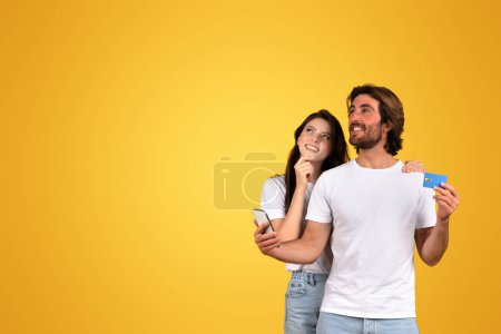 Photo for A european woman dreamily looking up and a bearded man smiling while holding a credit card and phone, both in white t-shirts, planning a purchase on a yellow background, studio - Royalty Free Image