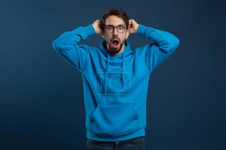Portrait of shocked scared young man with open mouth grabbing head and looking at camera in amazement, surprised terrified millennial guy standing against blue studio background, copy space