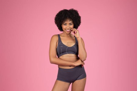 Photo for Smiling African American woman in gray top and shorts underwear confidently posing against pink backdrop and touching chin, smiling at camera - Royalty Free Image