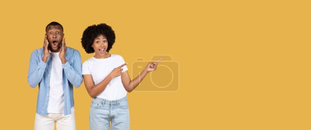 Téléchargez les photos : Couple afro-américain étonnant avec des expressions exagérées, l'homme avec les mains sur les joues et la femme pointant sur le côté, les deux sur un fond jaune moutarde uni, panorama - en image libre de droit