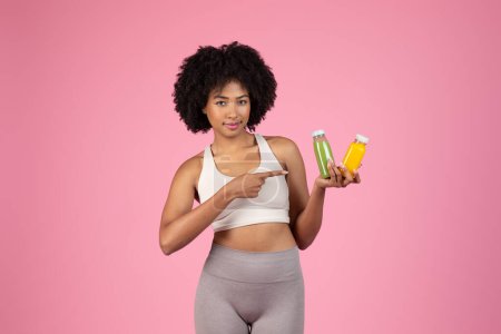 Téléchargez les photos : Convient aux jeunes femmes noires avec des cheveux bouclés naturels tenant et recommandant des bouteilles de jus de désintoxication verts et jaunes, en faisant un choix sur fond rose - en image libre de droit