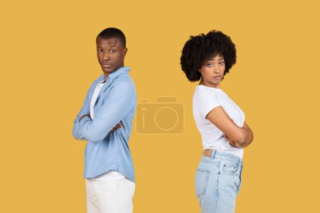 Téléchargez les photos : Un jeune homme et une jeune femme noirs en tenue décontractée se tiennent dos à dos avec les bras croisés, affichant des expressions de défi sur un fond jaune, studio. Problèmes relationnels - en image libre de droit