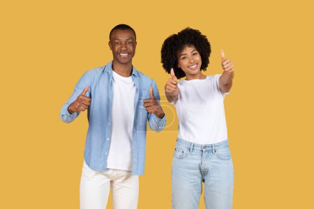 Téléchargez les photos : Heureux couple afro-américain donnant les pouces vers le haut, avec l'homme dans une chemise bleue et un pantalon blanc et la femme dans une chemise blanche et un jean, les deux souriant sur un fond jaune - en image libre de droit