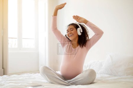 Foto de Mujer embarazada negra alegre escuchando música mientras se relaja en la cama en casa, madre embarazada afroamericana feliz en auriculares inalámbricos cantando y bailando mientras disfruta de la lista de reproducción favorita - Imagen libre de derechos