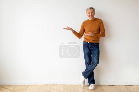 Photo for Handsome elderly man grandfather pointing at copy space over blank white wall in empty apartment, showing great offer, advertisement, full length. Relocation, moving, mortgage - Royalty Free Image