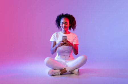 Téléchargez les photos : Joyeuse femme noire aux cheveux bouclés, portant du blanc, utilisant un smartphone tout en étant assise jambes croisées sur le sol avec un fond dégradé rose tendre à violet - en image libre de droit