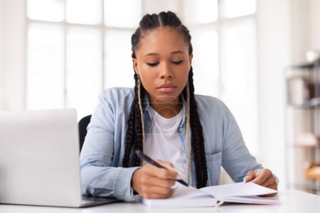Téléchargez les photos : Une jeune femme concentrée écrit intensément dans son carnet, avec un ordinateur portable ouvert devant elle, illustrant son dévouement à ses études tout en apprenant en ligne depuis la maison - en image libre de droit