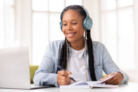 Photo for Joyful black female student with braids wearing headphones while studying and writing notes sitting in front of laptop in sunlit room at home interior - Royalty Free Image