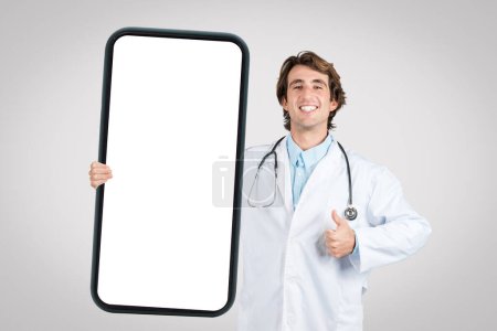 Joyful male doctor with stethoscope giving thumbs up holding giant smartphone frame with blank screen for app promotion or telemedicine concept