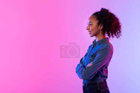 Photo for Side profile of self-assured black lady with curly hair, crossed arms, wearing denim jacket against vibrant pink gradient background, embodying confidence and style, copy space - Royalty Free Image