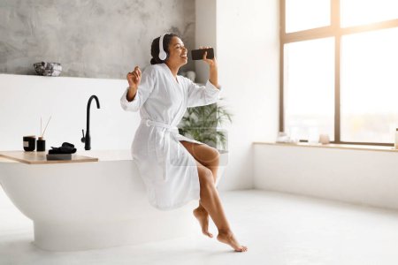 Photo for Cheerful black woman in bathrobe singing joyfully with smartphone in bathroom, happy african american female wearing headphones, embodying carefree relaxation and entertainment, copy space - Royalty Free Image