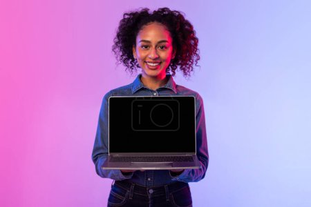 Photo for Confident black lady with curly hair and denim jacket holds an open laptop with blank screen, standing against vibrant pink and purple gradient background, mockup - Royalty Free Image