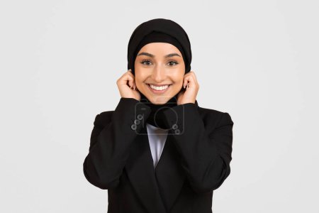 Téléchargez les photos : Radieuse jeune femme d'affaires en tenue professionnelle ajustant son hijab et regardant la caméra avec une expression joyeuse, isolée sur fond gris clair - en image libre de droit
