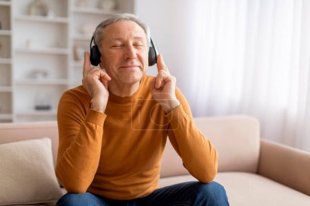 Téléchargez les photos : Homme âgé joyeux appréciant la bonne musique à la maison, assis sur le canapé avec les yeux fermés, en utilisant des écouteurs sans fil, écouter des chansons préférées, copier l'espace. Divertissement domestique - en image libre de droit
