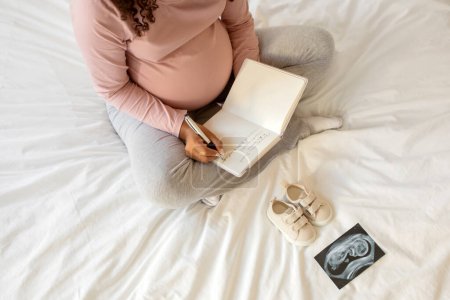 Téléchargez les photos : Femme enceinte écrivant des pensées dans son carnet de notes à la maison, assise sur le lit entourée de chaussures minuscules et d'une image d'échographie, future mère créant des souvenirs pendant sa grossesse ou faisant une liste de contrôle - en image libre de droit