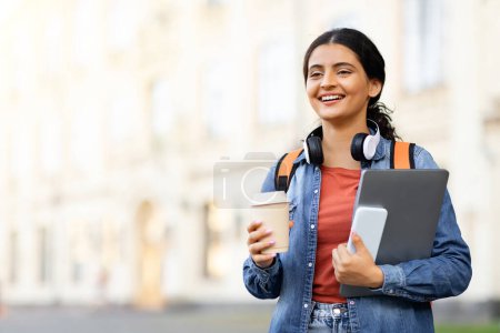 Photo for Positive pretty young eastern woman student with takeaway coffee, laptop and smartphone in her hands walking by university campus, looking at copy space. Online education concept - Royalty Free Image