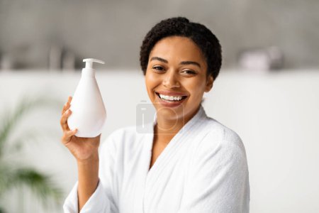 Photo for Beautiful Smiling African American Woman In Bathrobe Holding Bottle With Moisturising Body Lotion, Happy Black Lady Using New Skincare Product After Bath, Standing In Modern Bathroom Interior - Royalty Free Image