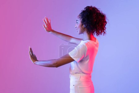 Téléchargez les photos : Joyeux cheveux bouclés dame noire geste ludique avec ses mains levées, montrant l'espace vide sur fond dégradé rose et violet bicolore - en image libre de droit