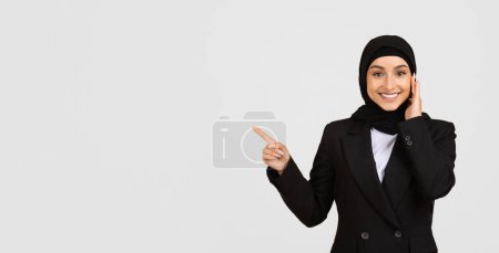 Photo for Cheerful muslim businesswoman wearing hijab confidently points aside at empty space, presenting space perfect for advertising content, panorama - Royalty Free Image