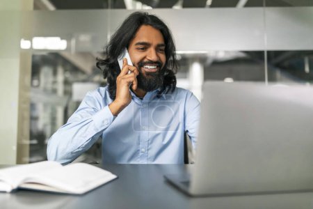 Photo for Eastern guy marketing director have phone call with client, office interior. Young indian man manager wearing formal outfit sitting at desk, talking on smartphone and looking at laptop screen - Royalty Free Image