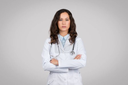 Portrait of confident woman doctor with folded arms, looking at camera, standing against grey background, showcasing professional healthcare personality