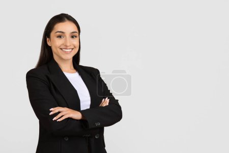 Photo for Smiling confident young professional woman with dark hair, wearing black suit and white shirt, standing with arms crossed on light background, copy space - Royalty Free Image