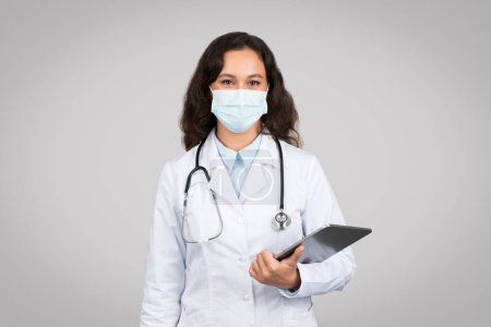 Foto de Joven terapeuta doctora en bata blanca, máscara protectora sosteniendo tableta, posando aislada sobre fondo gris del estudio. Asistencia sanitaria, consulta a distancia - Imagen libre de derechos