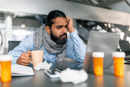 Photo for Exhausted sick young indian guy employee with warm scarf around his neck working on laptop at modern office, holding mug with hot drink, suffering from fever, have cold or flu - Royalty Free Image