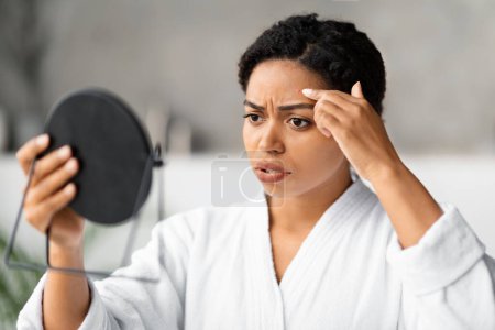 Téléchargez les photos : Femme noire préoccupée regardant dans un miroir à main au bouton sur son front, femme afro-américaine inquiète portant un peignoir repérant problème de peau tout en faisant la routine de beauté dans la salle de bain moderne - en image libre de droit
