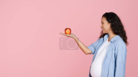 Foto de Sonriendo hermosa mujer embarazada joven mirando manzana orgánica roja en su palma, hambrienta esperando dama tienen bocadillo saludable, aislado en el fondo del estudio rosa, panorama con espacio para copiar - Imagen libre de derechos