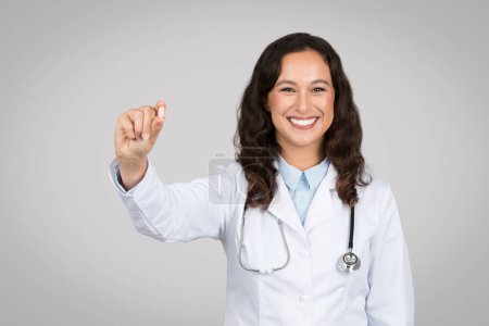 Glad lady doctor showing pill capsule, recommending treatment and smiling at camera, posing on gray background. Medical help, painkiller, vitamins and antidepressant