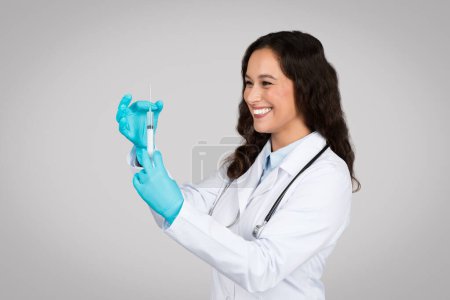 Photo for Cheerful young female doctor in white lab coat and gloves carefully preparing syringe for vaccination, representing medical procedures and patient care - Royalty Free Image