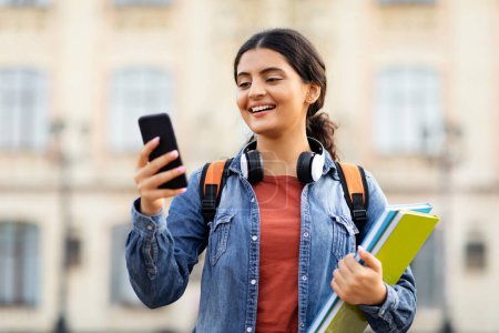 Photo for Online learning, e-education concept. Happy young indian lady student using phone outdoors, carrying backpack and textbooks, going to university, checking her schedule online - Royalty Free Image