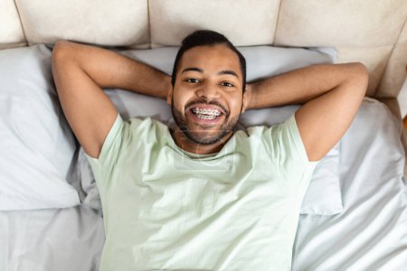 Téléchargez les photos : Vue ci-dessus de joyeux jeune homme afro-américain barbu gai allongé dans le lit avec les bras derrière la tête, souriant à la caméra, rêvant d'un jour de chance prospère, se sentant bien reposé - en image libre de droit