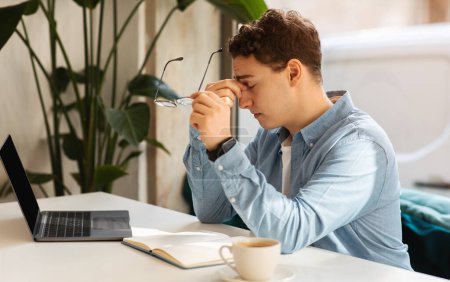 Téléchargez les photos : Un jeune homme caucasien triste et fatigué se sentant stressé ou fatigué en travaillant, tenant ses lunettes d'une main et se frottant les yeux, assis à un bureau avec un ordinateur portable et une tasse de café - en image libre de droit