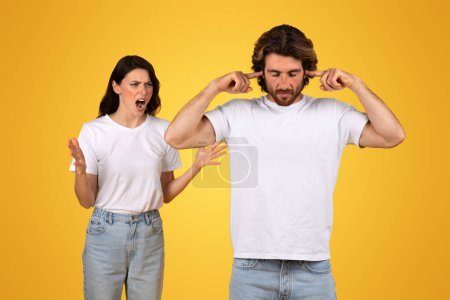 Annoyed angry caucasian woman yelling with hands open in disbelief while man beside her plugs his ears, both wearing white t-shirts, against a bright yellow background, studio