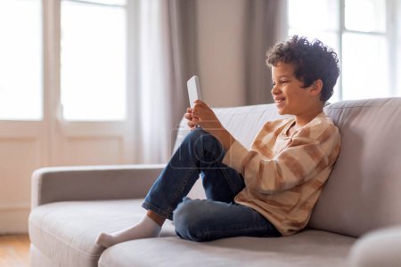 Photo for Cheerful black boy sitting relaxed on sofa, deeply engaged in his smartphone, happy african american male kid using mobile phone, playing game or browsing internet, relaxing on couch at home - Royalty Free Image